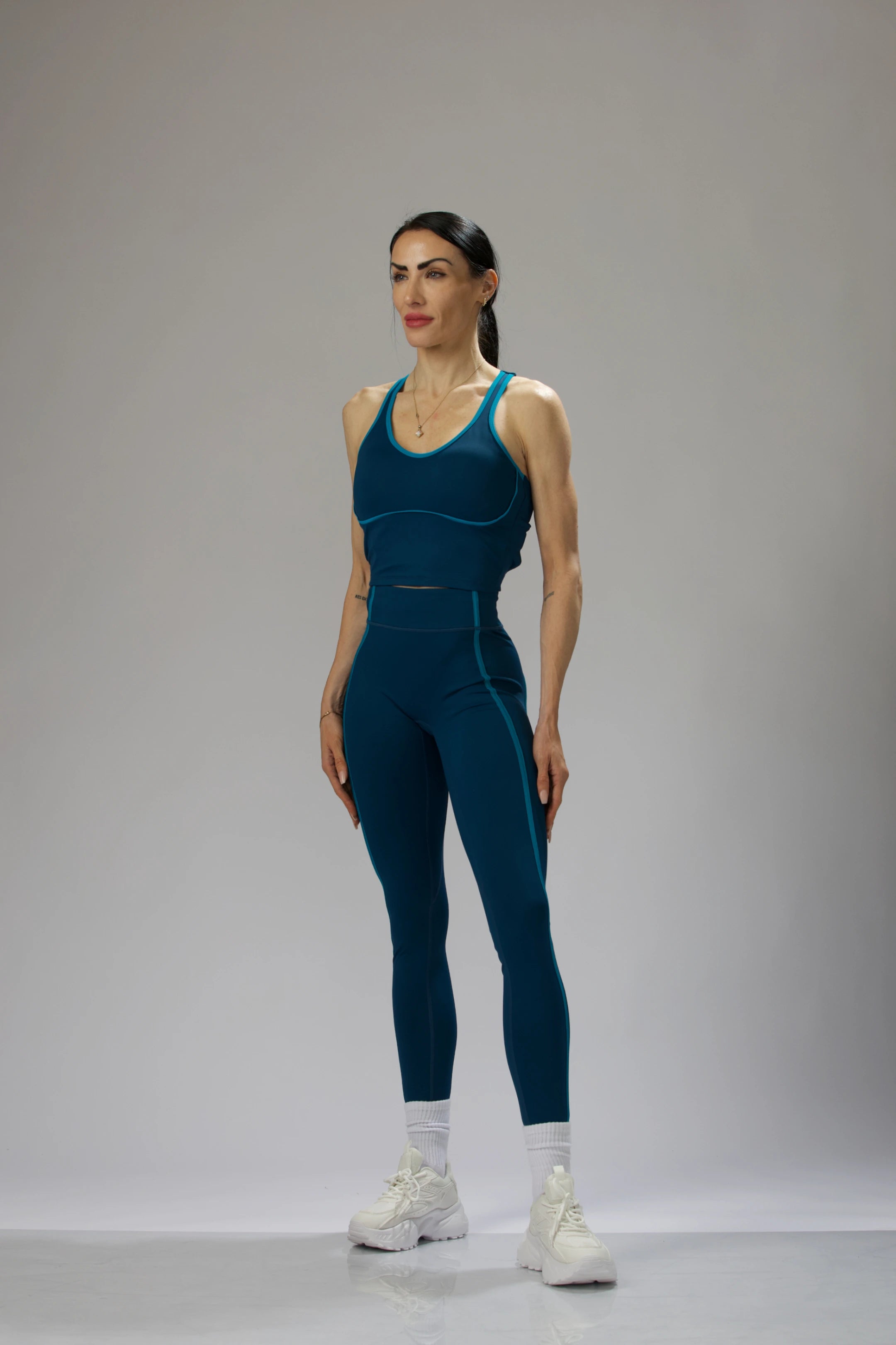 A female model in a bright teal sports bra and leggings, standing with one hand on her hip.
