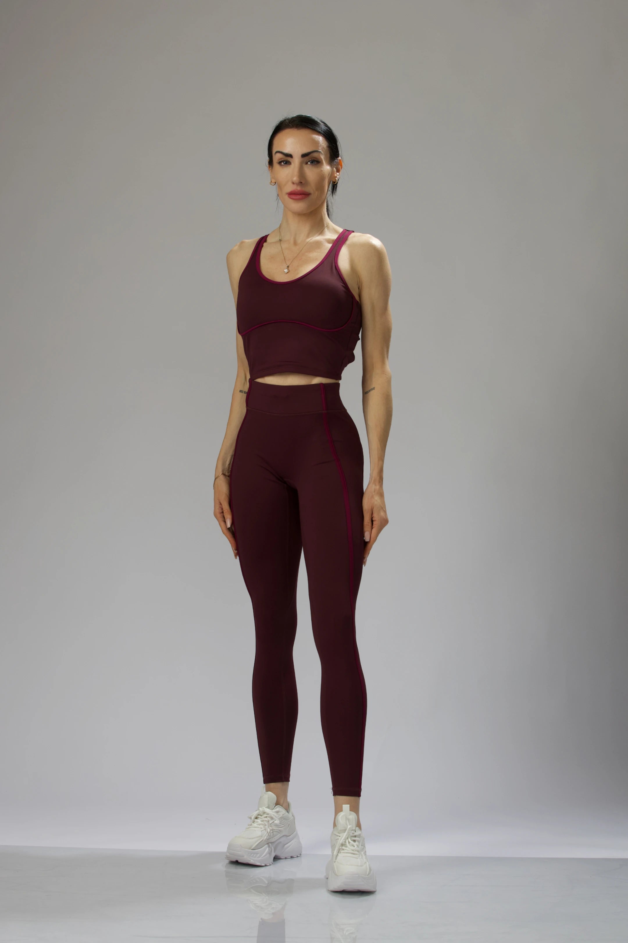 A female model in a dark wine-colored sports bra and leggings, posing in a studio setting.
