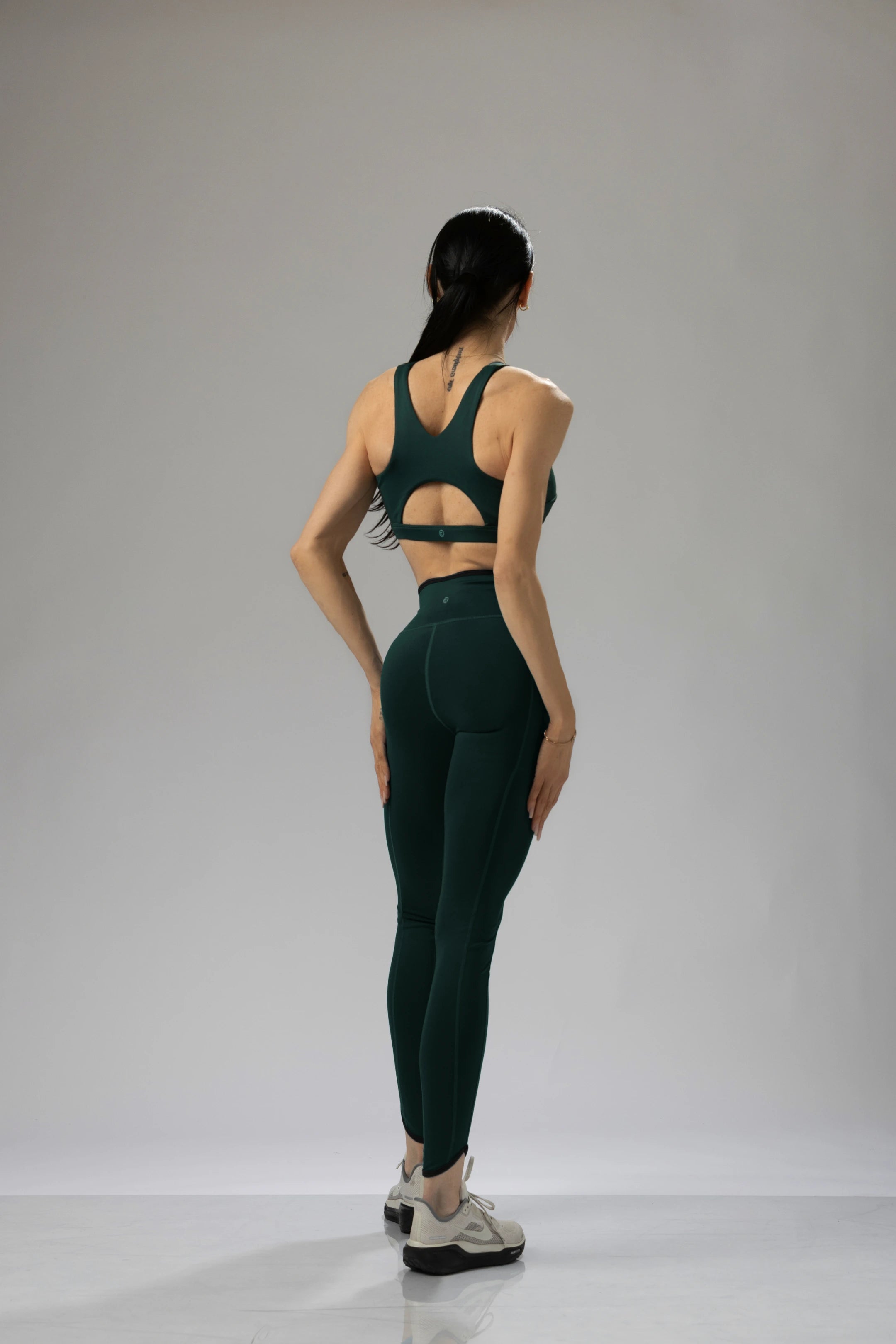 Rear view of a woman in a dark green sports bra and leggings, showcasing the unique back cut-out.
